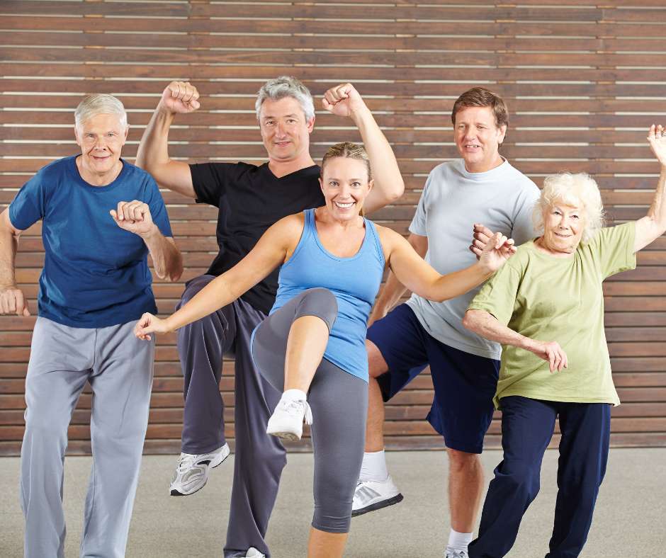 Eine Gruppe von fünf Menschen unterschiedlichen Alters, von jung bis alt, führt gemeinsam eine Fitnessübung durch. Sie tragen bequeme Sportkleidung und lächeln, während sie die Übung machen. Der Hintergrund besteht aus einer Holzwand. Das Bild vermittelt ein Gefühl von Gemeinschaft und zeigt, dass Fitness für alle Altersgruppen geeignet ist. Es betont die Inklusivität und den Spaß am Training in einem Fitnessstudio.