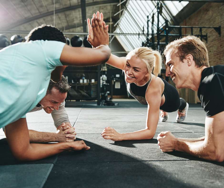 Eine Gruppe von vier Menschen trainiert gemeinsam in einem Fitnessstudio und führt Plank-Übungen aus. Zwei von ihnen geben sich in der Mitte des Bildes ein High-Five, während sie lachen und sich gegenseitig motivieren. Die Atmosphäre ist energiegeladen und fröhlich, und im Hintergrund sind Fitnessgeräte und Hanteln zu sehen. Das Bild vermittelt Gemeinschaftsgefühl, Spaß am Training und die positive Energie, die durch Gruppentraining entstehen kann.