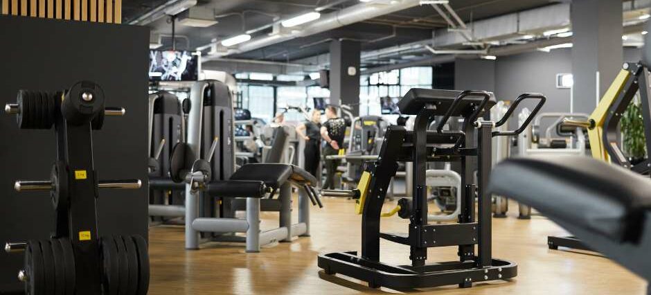 Ein modernes Fitnessstudio mit einer Vielzahl von Fitnessgeräten und Hantelbänken. Der Raum ist gut beleuchtet und verfügt über eine stilvolle Inneneinrichtung mit Holz- und Metallakzenten. Im Hintergrund sind einige Mitglieder zu sehen, die an den Geräten trainieren. Das Bild vermittelt ein Gefühl von Professionalität und gut ausgestatteten Trainingsmöglichkeiten, ideal für eine Social Media Strategie, die die hochwertigen Einrichtungen und die einladende Atmosphäre des Fitnessstudios hervorhebt.